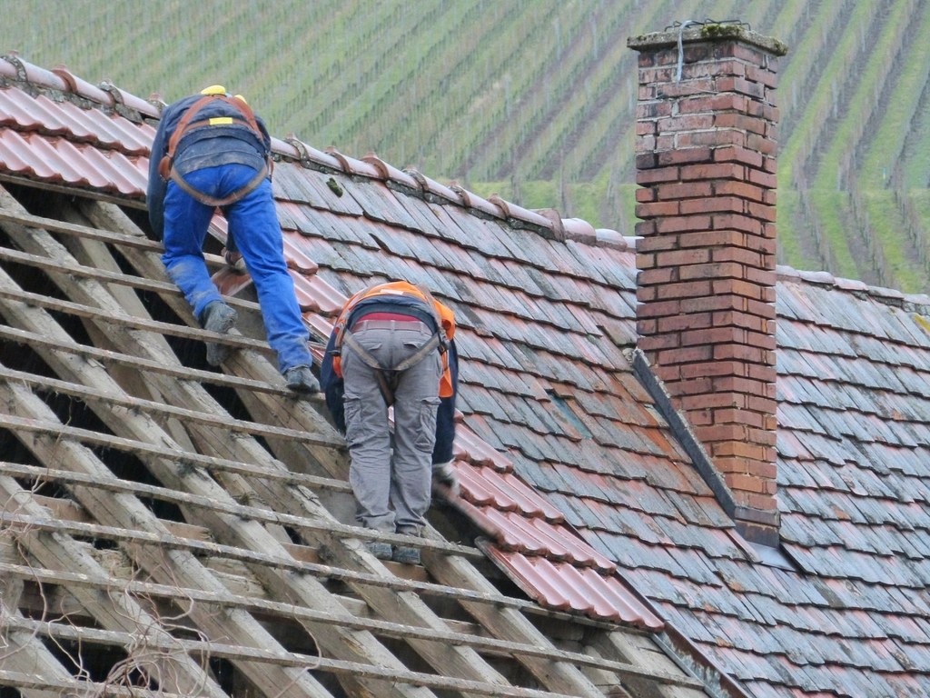 Two roofers on the battens re-tiling a pitched roof beside a brick chimney